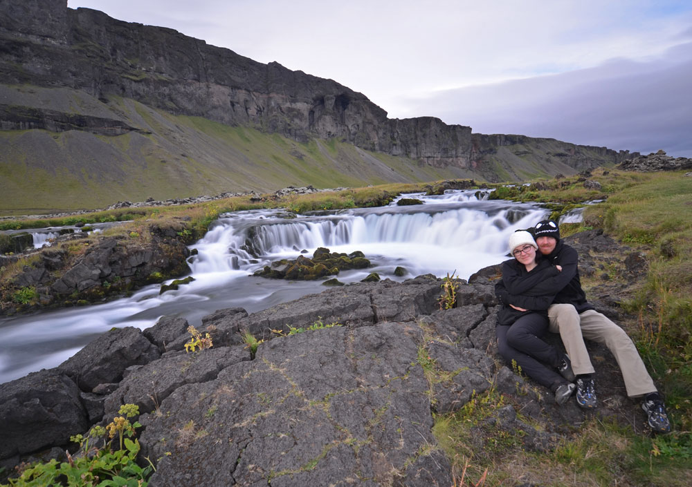Brooke and Buddy sitting next to a road-side waterfall during their Iceland Vacation