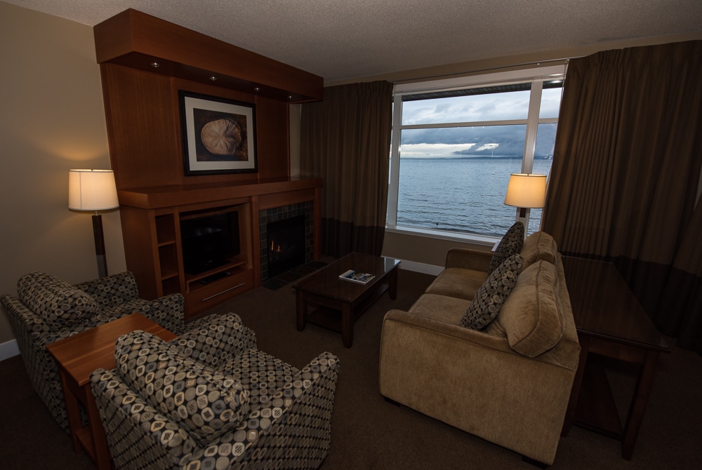 Living room with TV, fireplace, plenty of seating and large open windows looking out over the water.