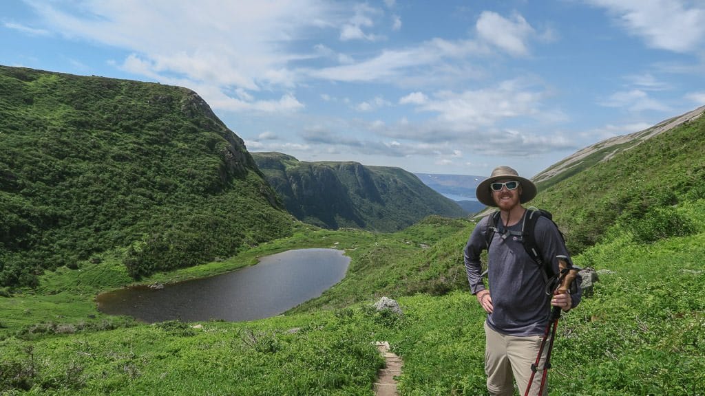 Buddy posing next to the narrow trail surrounded by grass near a beautiful lake in Gros Morne National Park