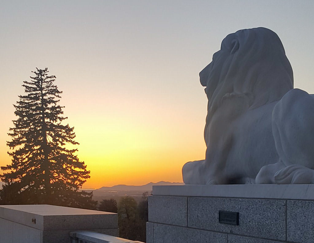 Sunset at the Utah Capitol Building. Lion statue in the foreground, and a tree and mountains in the background.