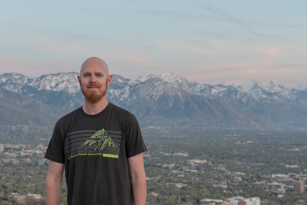 Buddy on the top of Ensign Hill with the mountains behind him and he is wearing a shirt with a mountain on it.