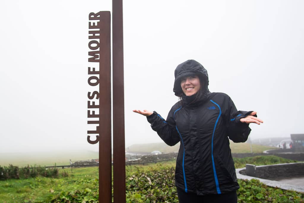 Brooke in her rain gear standing next to the Cliffs of Moher sign with no visibility behind her due to the thick fog