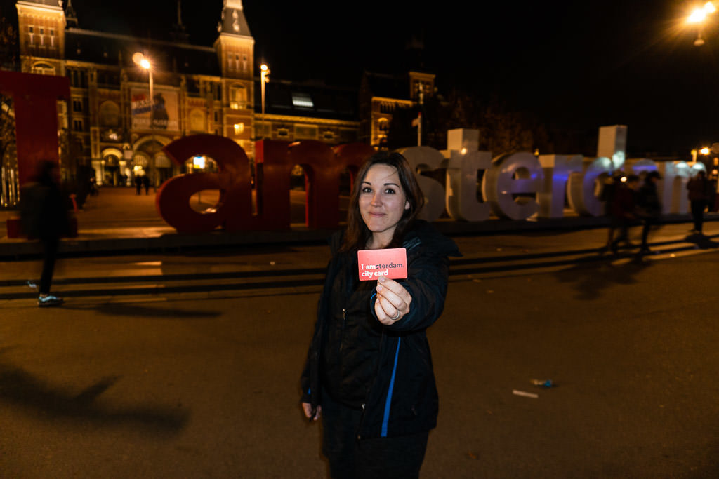 Brooke holding the i amsterdam city card in front of I amsterdam sign at night